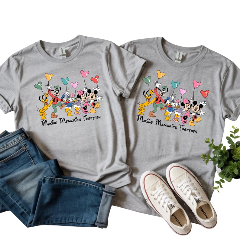 May include: Two gray t-shirts with a colorful graphic of Disney characters holding balloons. The text "Making Memories Together" is below the graphic. Blue jeans and white sneakers are next to the shirts.