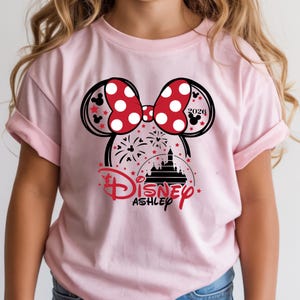 May include: Pale pink t-shirt with a Minnie Mouse graphic. The design includes a red polka-dot bow, mouse ears, fireworks, a castle, and the word "Disney" in red script. The name "Ashley" is below the Disney logo.
