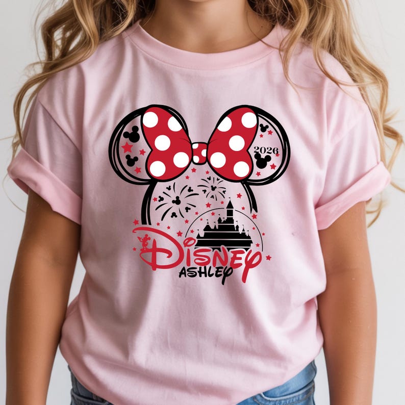 May include: Pale pink t-shirt with a Minnie Mouse graphic. The design includes a red polka-dot bow, mouse ears, fireworks, a castle, and the word "Disney" in red script. The name "Ashley" is below the Disney logo.