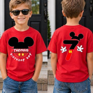 May include: Red t-shirt with a Mickey Mouse design. The front of the shirt has the text "THOMAS BIRTHDAY BOY" and a Mickey Mouse head. The back of the shirt has a number 7 with Mickey Mouse gloves and head.