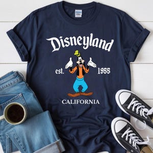Op de afbeelding: Marineblauw T-shirt met het Disneyland-logo, Goofy-cartoonfiguur en de tekst "est. 1955 CALIFORNIA". Het shirt wordt getoond met een blauwe jeans en zwarte sneakers.