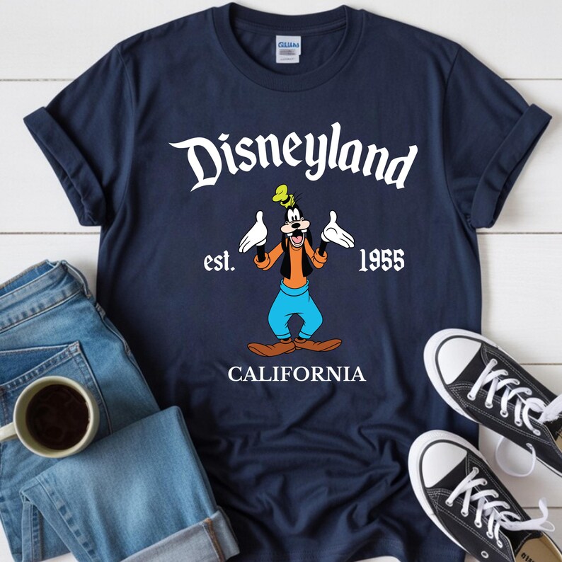 Op de afbeelding: Marineblauw T-shirt met het Disneyland-logo, Goofy-cartoonfiguur en de tekst "est. 1955 CALIFORNIA". Het shirt wordt getoond met een blauwe jeans en zwarte sneakers.