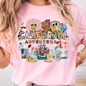 May include: A pale pink sweatshirt with a vibrant graphic. The design features the words "CALIFORNIA ADVENTURE" alongside cartoon characters and amusement park elements, including Mickey Mouse and a Ferris wheel.