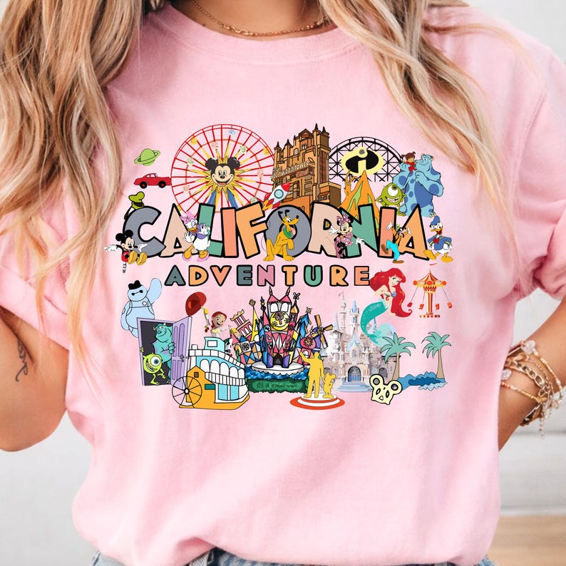 May include: A pale pink sweatshirt with a vibrant graphic. The design features the words "CALIFORNIA ADVENTURE" alongside cartoon characters and amusement park elements, including Mickey Mouse and a Ferris wheel.