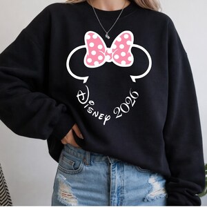 May include: Black sweatshirt featuring a white Minnie Mouse design with a pink polka-dot bow and the text "Disney 2026". The design includes the iconic mouse ears and a cursive font.