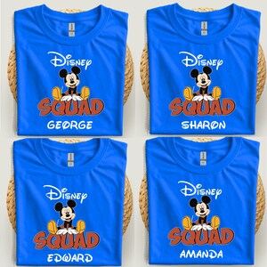 Op de afbeelding: Vier blauwe t-shirts met het Disney-logo, een Mickey Mouse-afbeelding en het woord "SQUAD" in oranje en rood. Elk shirt heeft een andere naam: George, Sharon, Edward en Amanda. De shirts worden gepresenteerd op een geweven oppervlak.
