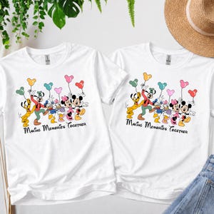 May include: Two white t-shirts with a colorful graphic of Disney characters holding balloon bouquets. The text "Making Memories Together" is printed below the characters. The shirts are casual wear.