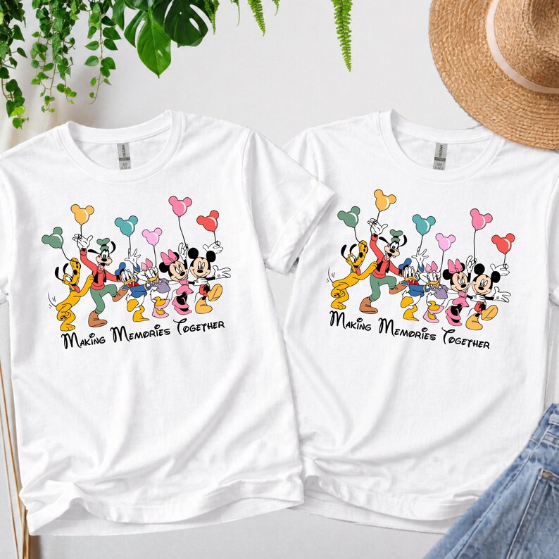 May include: Two white t-shirts with a colorful graphic of Disney characters holding balloon bouquets. The text "Making Memories Together" is printed below the characters. The shirts are casual wear.