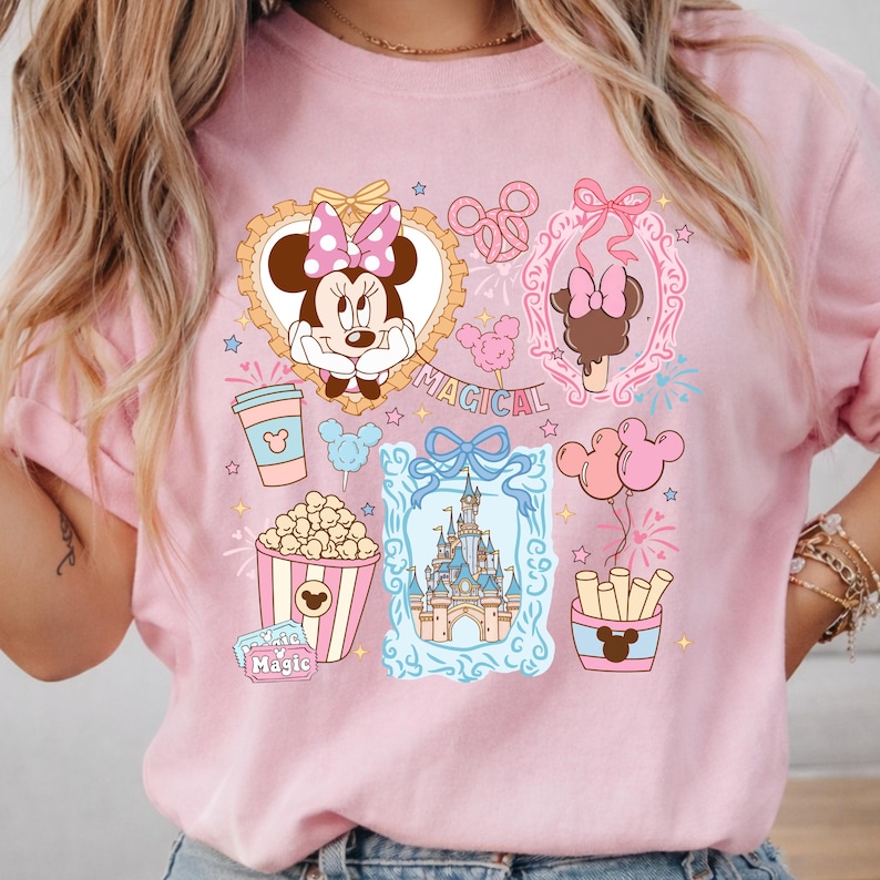 Puede incluir: Camiseta rosa con un dise&ntilde;o gr&aacute;fico colorido que presenta a Minnie Mouse, un castillo, palomitas de ma&iacute;z y otros elementos de parques tem&aacute;ticos. La palabra "Magical" est&aacute; incluida. La camiseta tiene cuello redondo y mangas cortas.