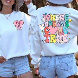 May include: White sweatshirt with a colourful graphic on the front and back. The front features a pink bow and castle design. The back displays the text "WHERE DREAMS COME TRUE" in a retro font with Mickey Mouse-themed accents.