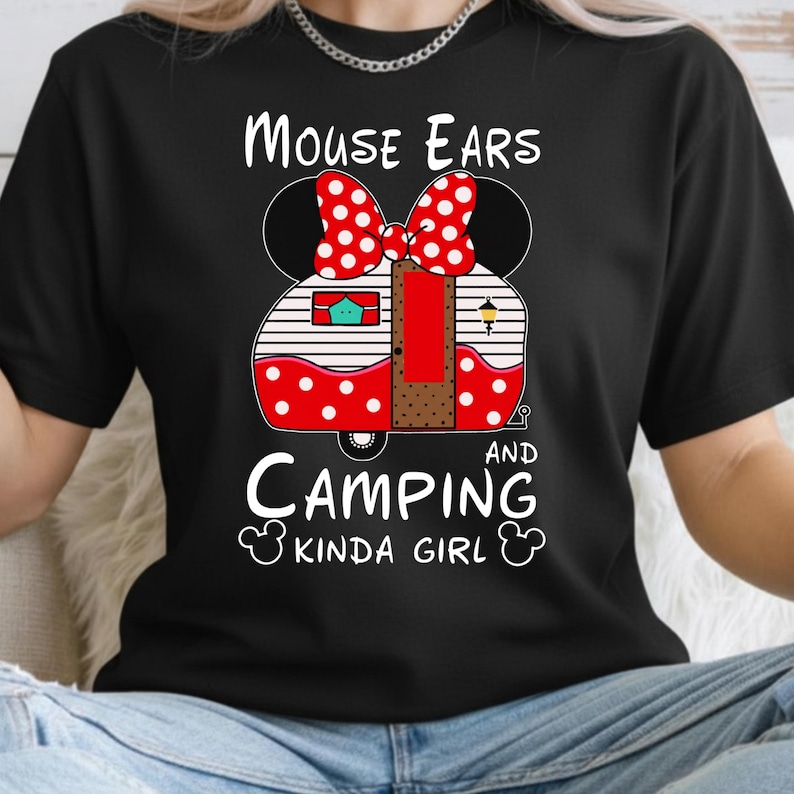 Puede incluir: Camiseta negra con un dise&ntilde;o de caravana con forma de orejas de rat&oacute;n, un lazo rojo de lunares y las palabras "MOUSE EARS AND CAMPING KINDA GIRL" en escritura blanca.