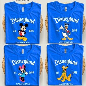 Op de afbeelding: Vier blauwe T-shirts met het Disneyland-logo en tekenfilmfiguren: Mickey Mouse, Donald Duck, Minnie Mouse en Pluto. Elk shirt heeft de tekst "est. 1955 CALIFORNIA".
