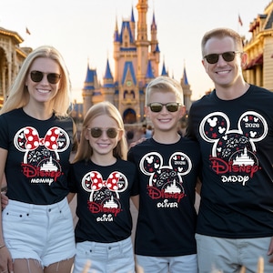 May include: Four people wearing black Disney t-shirts with a Mickey Mouse head design. The design includes a red and white bow, fireworks, and the text "Disney" and "2026". The shirts are personalized with "Mommy", "Olivia", "Oliver", and "Daddy".