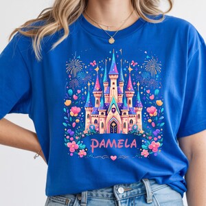 K&ouml;nnte beinhalten: K&ouml;nigsblaues T-Shirt mit einem farbenfrohen Schlossdesign, Feuerwerk und floralen Elementen. Der Name "PAMELA" ist in einer dekorativen Schriftart geschrieben. Das Schlossdesign hat rosa, blaue und violette T&uuml;rme, umgeben von Blumen.