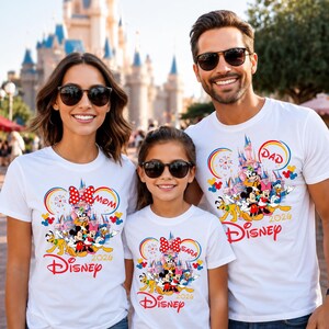 Op de afbeelding: Drie mensen dragen witte t-shirts met een Disney-thema. Elk shirt heeft een kleurrijke afbeelding van Disney-personages, een kasteel en het woord "Disney" in het rood. De shirts hebben ook de woorden "Mom", "Dad" en "Sara".
