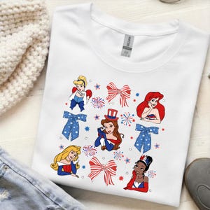 May include: White t-shirt with cartoon princesses, red and blue bows, fireworks, and stars. The princesses are in red, white, and blue outfits. The shirt is made of a soft material.