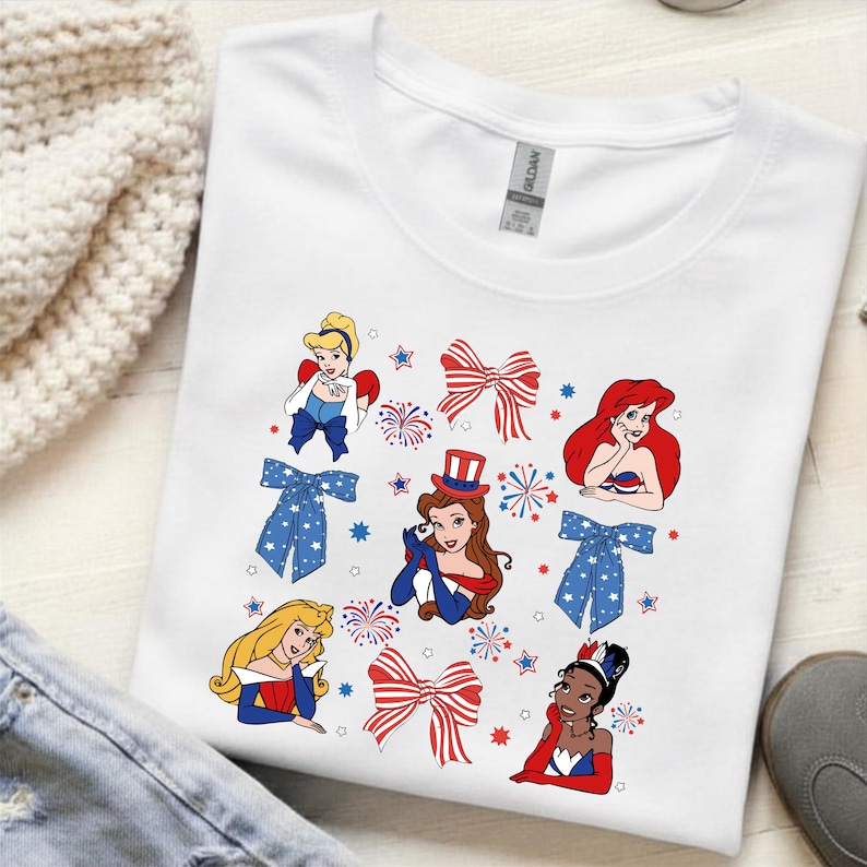 May include: White t-shirt with cartoon princesses, red and blue bows, fireworks, and stars. The princesses are in red, white, and blue outfits. The shirt is made of a soft material.