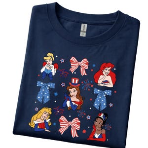 May include: Dark blue t-shirt with cartoon princess illustrations and patriotic bow designs. The shirt has a crew neck and short sleeves. The graphic includes red, white, and blue colours, with star and firework accents.
