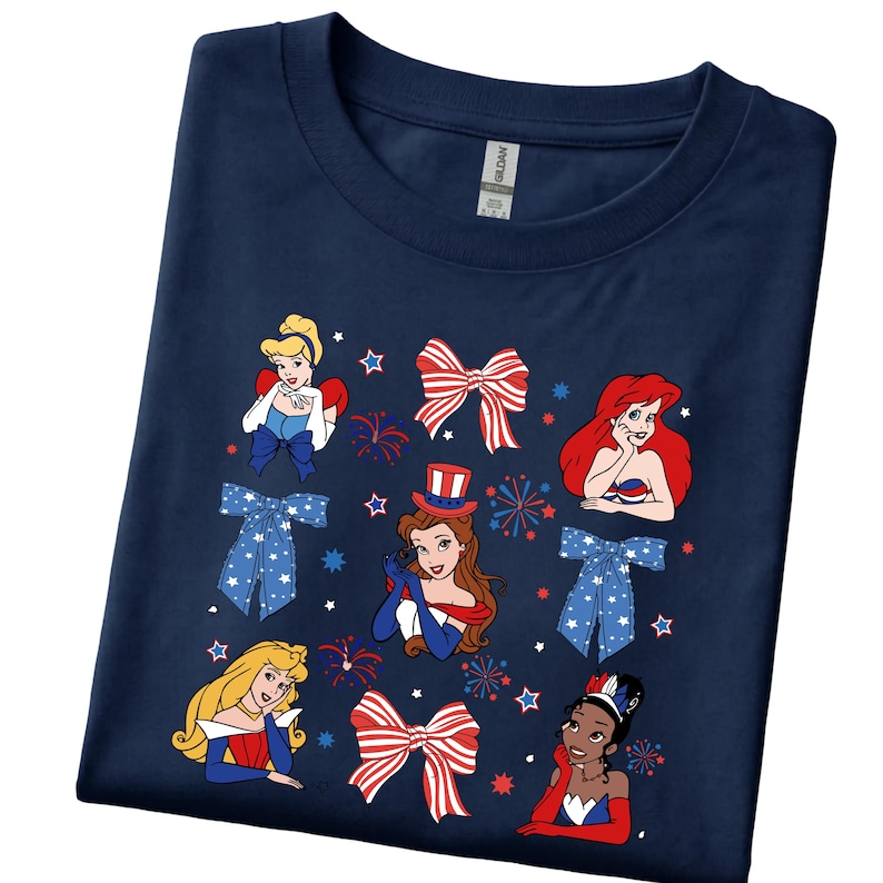 May include: Dark blue t-shirt with cartoon princess illustrations and patriotic bow designs. The shirt has a crew neck and short sleeves. The graphic includes red, white, and blue colours, with star and firework accents.