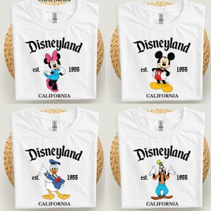 Op de afbeelding: Vier witte t-shirts met Disneyland-ontwerpen. Elk shirt toont het Disneyland-logo, het jaar 1955 en het woord "CALIFORNIA". De shirts tonen Minnie Mouse, Mickey Mouse, Donald Duck en Goofy.