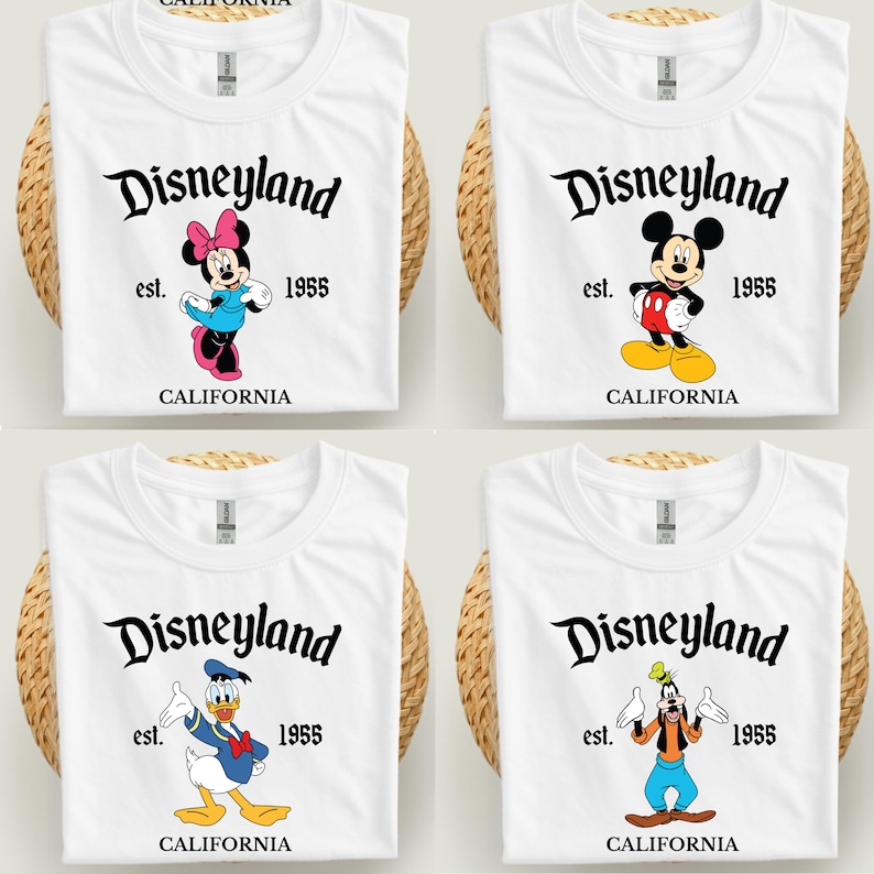 Op de afbeelding: Vier witte t-shirts met Disneyland-ontwerpen. Elk shirt toont het Disneyland-logo, het jaar 1955 en het woord "CALIFORNIA". De shirts tonen Minnie Mouse, Mickey Mouse, Donald Duck en Goofy.