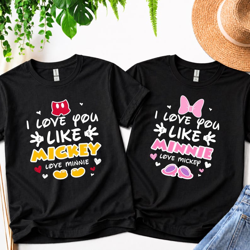 Pode incluir: Duas camisetas pretas com texto branco. Uma diz "I love you like Mickey" com la&ccedil;o vermelho e texto amarelo. A outra diz "I love you like Minnie" com la&ccedil;o rosa e texto rosa. Ambas t&ecirc;m cora&ccedil;&otilde;es.