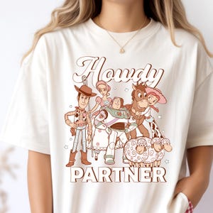May include: Cream-colored t-shirt featuring a cartoon graphic of characters from a popular animated film. The design includes the word "Howdy" above the word "Partner". The characters are in shades of brown, pink, green, and white.