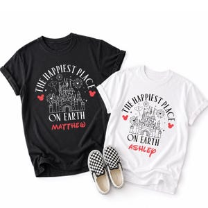 May include: Two t-shirts, one black and one white, with a castle graphic and the text "The Happiest Place on Earth." The black shirt has the name "Matthew" and the white shirt has the name "Ashley." Checkered slip-on shoes are in front.