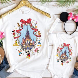 May include: White t-shirts and a onesie featuring a colorful illustration of a castle within a red ribbon frame, surrounded by cartoon characters. The shirts are hanging on a gold hanger, with a Minnie Mouse ear headband with a pink bow.