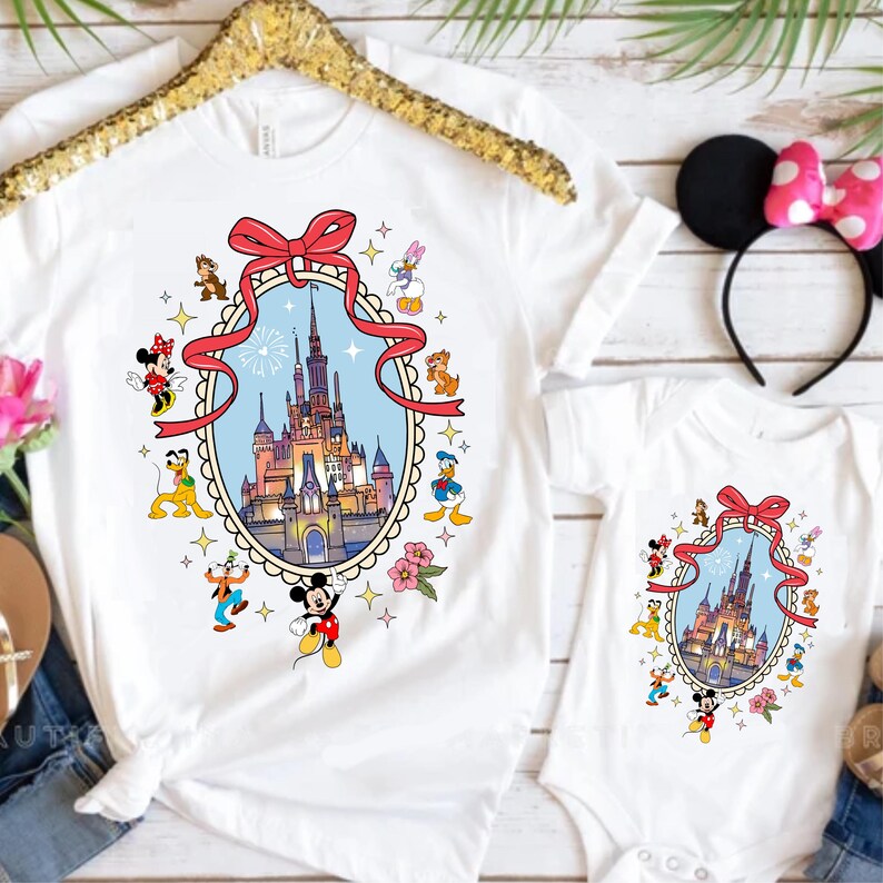May include: White t-shirts and a onesie featuring a colorful illustration of a castle within a red ribbon frame, surrounded by cartoon characters. The shirts are hanging on a gold hanger, with a Minnie Mouse ear headband with a pink bow.