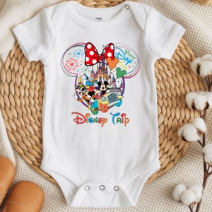 Puede incluir: Body de beb&eacute; blanco con un colorido gr&aacute;fico de Disney Trip. El dise&ntilde;o presenta a Mickey y Minnie Mouse, el Pato Donald y Goofy en un coche, con un castillo y fuegos artificiales. Un lazo rojo con lunares en la parte superior.