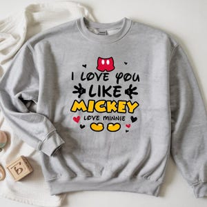 Pode incluir: Um moletom cinza claro com o texto "I love you like Mickey" em preto, amarelo e vermelho. O design inclui as orelhas, m&atilde;os e p&eacute;s do Mickey Mouse, junto com cora&ccedil;&otilde;es. O moletom &eacute; um estilo casual e confort&aacute;vel.