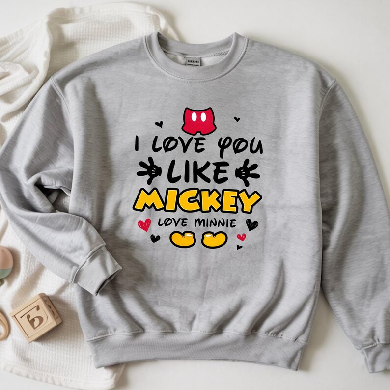 Pode incluir: Um moletom cinza claro com o texto "I love you like Mickey" em preto, amarelo e vermelho. O design inclui as orelhas, m&atilde;os e p&eacute;s do Mickey Mouse, junto com cora&ccedil;&otilde;es. O moletom &eacute; um estilo casual e confort&aacute;vel.
