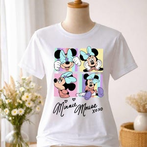 Pu&ograve; includere: T-shirt bianca con una grafica colorata di Minnie Mouse in quattro pose diverse, ciascuna su uno sfondo pastello. Le parole "Minnie Mouse xoxo" sono scritte in corsivo sotto. Minnie indossa un fiocco azzurro.