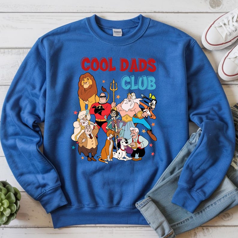 Pode incluir: Camisola azul com o texto "COOL DADS CLUB" em vermelho e azul. O gr&aacute;fico apresenta personagens de desenhos animados, incluindo um le&atilde;o, um super-her&oacute;i e outras figuras. A camisola &eacute; combinada com jeans.