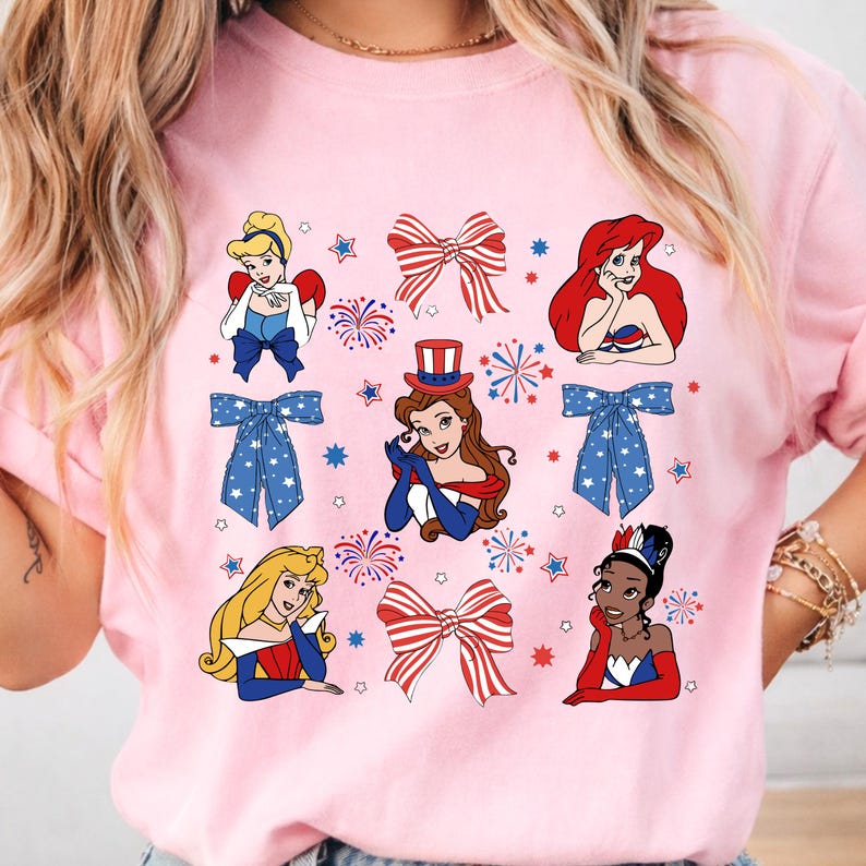 May include: A pink sweatshirt with cartoon illustrations of princesses, red, white, and blue bows, fireworks, and stars. The princesses are dressed in patriotic attire. A festive design for a themed celebration.