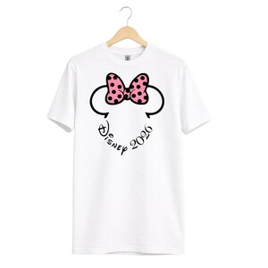 May include: White t-shirt featuring a Minnie Mouse design. The design includes a pink bow with black polka dots, black mouse ears, and the text "Disney 2026" in a script font.