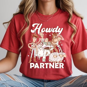 May include: Red t-shirt featuring a graphic of cartoon characters with the text "Howdy Partner" in white. The design includes a cowboy, space ranger, horse, and sheep. The shirt is a crew neck.