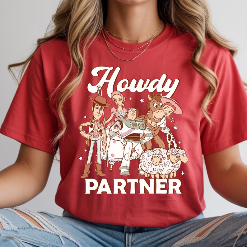 May include: Red t-shirt featuring a graphic of cartoon characters with the text "Howdy Partner" in white. The design includes a cowboy, space ranger, horse, and sheep. The shirt is a crew neck.