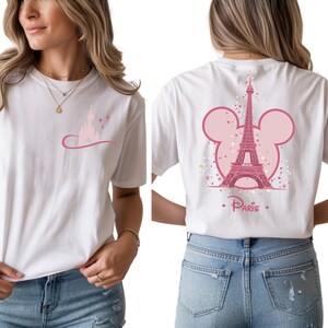 May include: White t-shirt featuring a pink graphic of a castle and a curved line with stars on the front. The back of the shirt has a pink Eiffel Tower design with a Mickey Mouse head outline and the word "Paris".