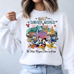 K&ouml;nnte beinhalten: Wei&szlig;es Sweatshirt mit einem farbenfrohen Aufdruck von Disney-Figuren und Wahrzeichen. Das Design zeigt Micky und Minnie Maus, Donald Duck und Goofy, mit dem Text "Walt Disney World" und "The Most Magical Place on Earth".