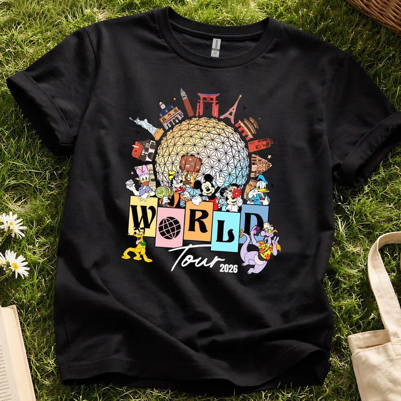 May include: Black t-shirt with a vibrant graphic of Disney characters and world landmarks. The design includes the text "WORLD Tour 2026" with characters such as Mickey Mouse and Donald Duck. The shirt is shown on a grassy background.
