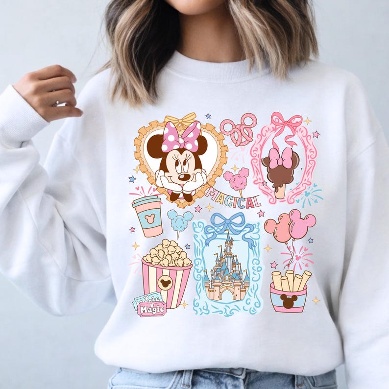 Puede incluir: Sudadera blanca con un dise&ntilde;o gr&aacute;fico colorido con Minnie Mouse, palomitas de ma&iacute;z, un castillo y la palabra "MAGICAL". El dise&ntilde;o incluye acentos rosas, azules y amarillos, con estrellas y otros elementos decorativos.