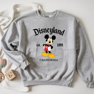 Op de afbeelding: Een lichtgrijs sweatshirt met het woord "Disneyland" in zwart schrift boven een Mickey Mouse-afbeelding. De afbeelding bevat de tekst "est. 1955" en "CALIFORNIA" onder het personage. De sweatshirt heeft lange mouwen.