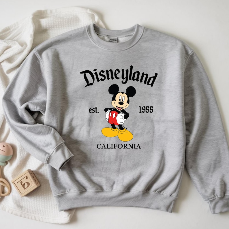 Op de afbeelding: Een lichtgrijs sweatshirt met het woord "Disneyland" in zwart schrift boven een Mickey Mouse-afbeelding. De afbeelding bevat de tekst "est. 1955" en "CALIFORNIA" onder het personage. De sweatshirt heeft lange mouwen.
