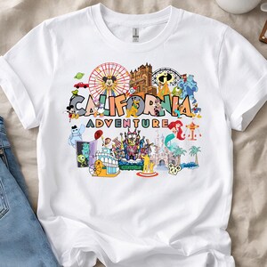 May include: White t-shirt with a vibrant graphic that reads "CALIFORNIA ADVENTURE". The design incorporates cartoon characters and landmarks, including a Ferris wheel, a castle, and a mermaid, in a colourful arrangement.