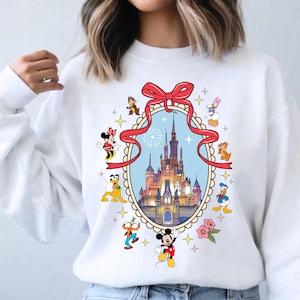 May include: White sweatshirt featuring a colorful illustration of a castle within a decorative frame, surrounded by cartoon characters like Mickey Mouse, Minnie Mouse, and Donald Duck. A red bow adorns the top of the frame. The design includes stars and flowers.