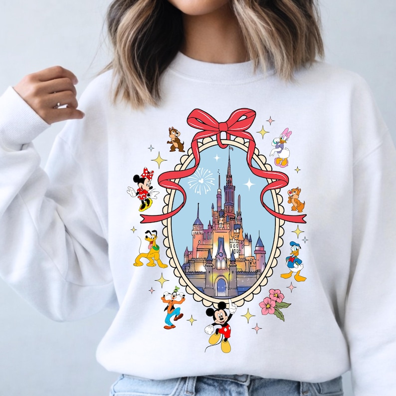 May include: White sweatshirt featuring a colorful illustration of a castle within a decorative frame, surrounded by cartoon characters like Mickey Mouse, Minnie Mouse, and Donald Duck. A red bow adorns the top of the frame. The design includes stars and flowers.