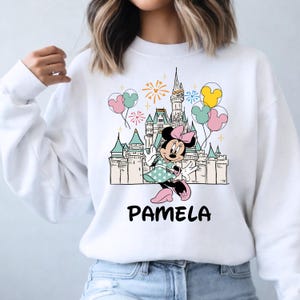 Może przedstawiać: Biała bluza z nadrukiem z kresk&oacute;wki Minnie Mouse w sukience w kropki, przed zamkiem z balonami i fajerwerkami. Imię "PAMELA" jest wydrukowane czarnymi literami poniżej.