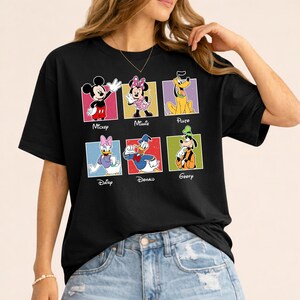 May include: Black t-shirt featuring six colorful squares with Disney characters: Mickey, Minnie, Pluto, Daisy, Donald, and Goofy. Each character is labeled with their name in a bold, white font. The shirt is worn by a person.